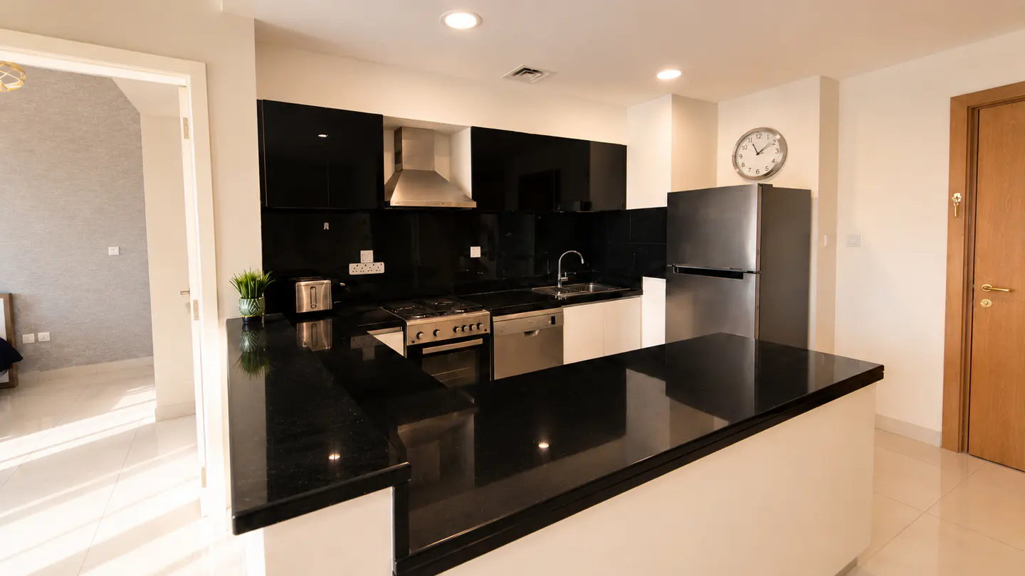 A modern kitchen with black cabinets, stainless steel appliances, and a black granite countertop island. A doorway leads to a bedroom.