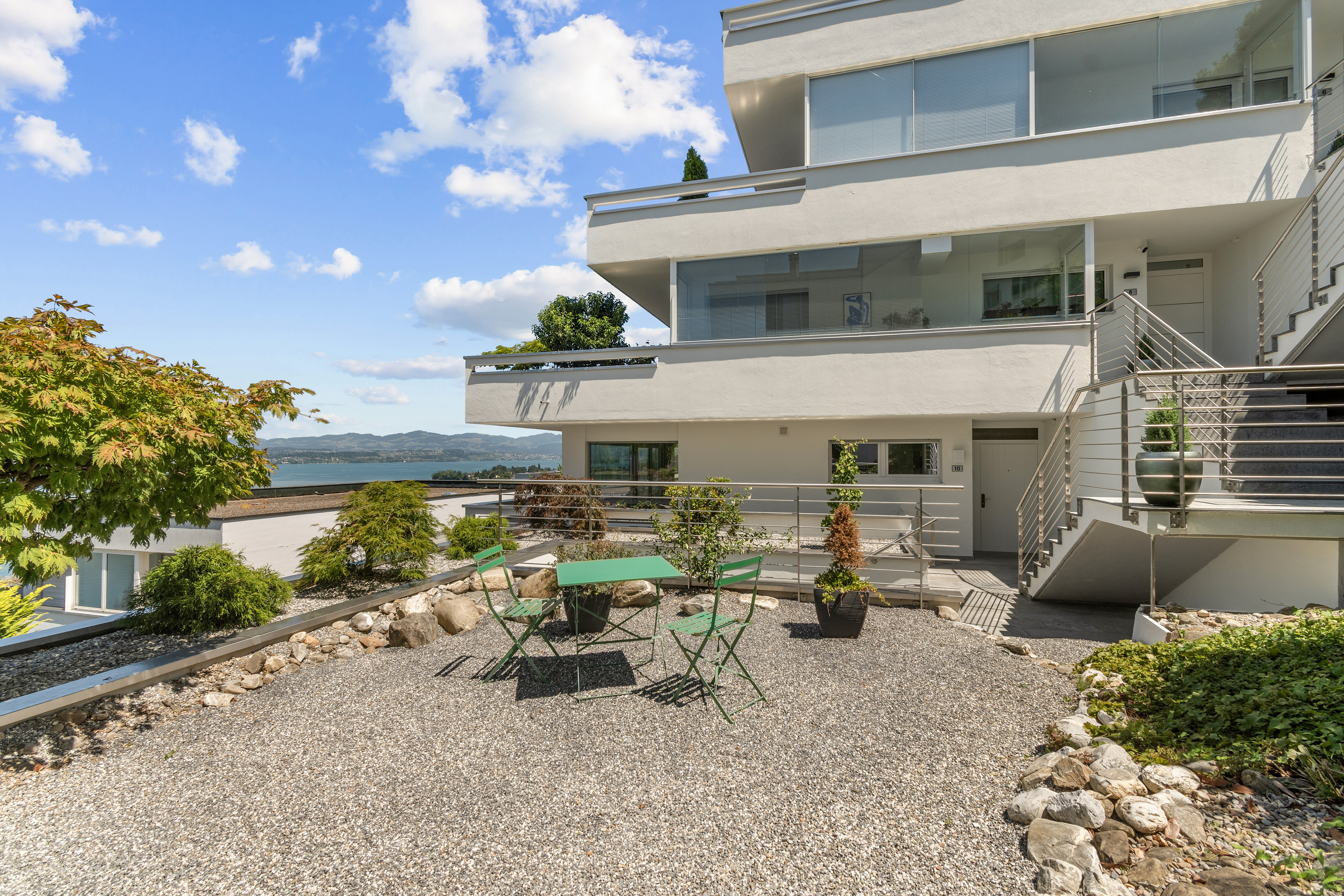 Modern white multi-story home with a gravel patio, green bistro set, and scenic lake view under a blue sky.