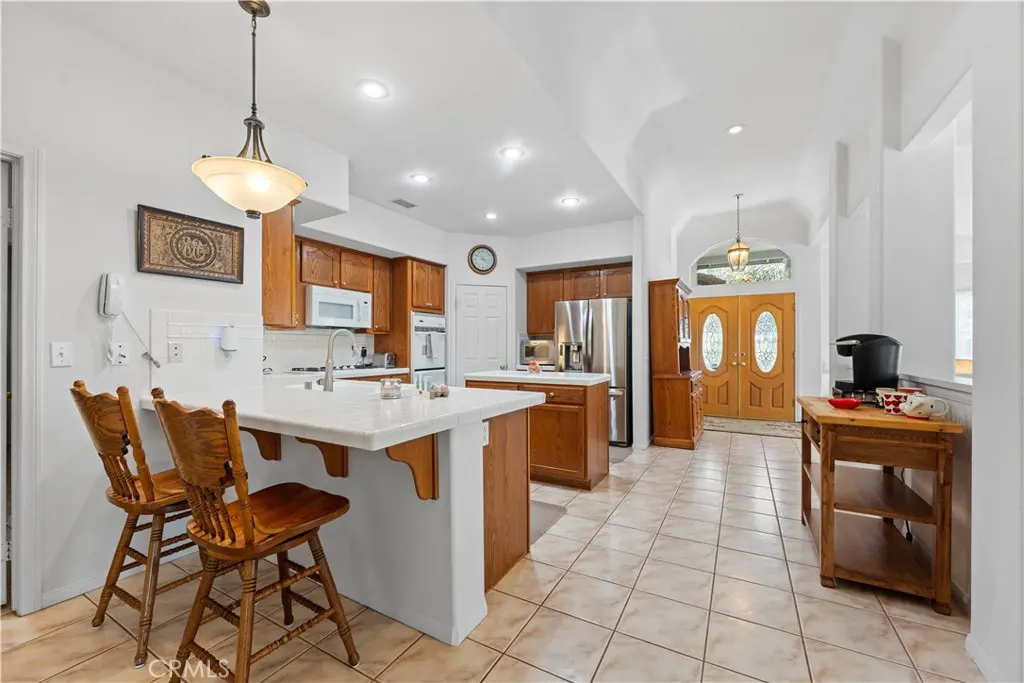A bright kitchen with wood cabinets, white countertops, and tile floors leads to a wood double door entrance.