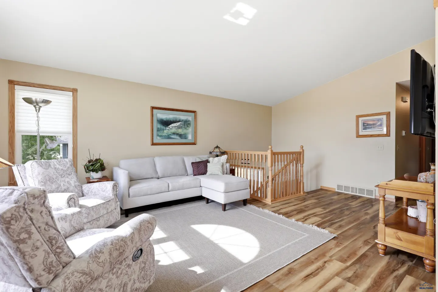 Living room with beige walls, wood-look floors, a white sectional, patterned armchairs, and a wooden staircase railing.
