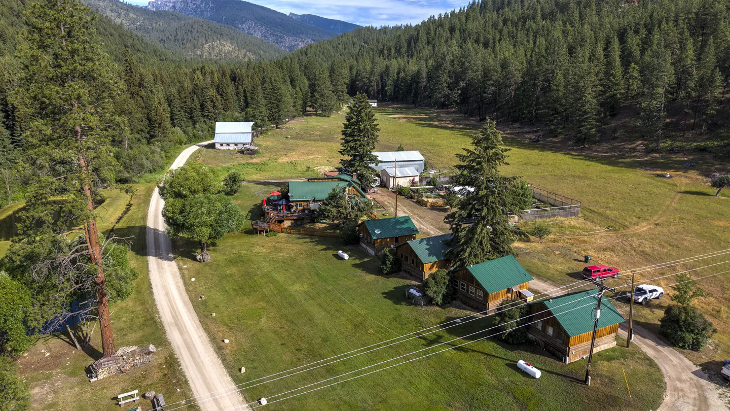 Aerial view of a rural property with cabins, green roofs, and a dirt road, surrounded by a forest and mountains.