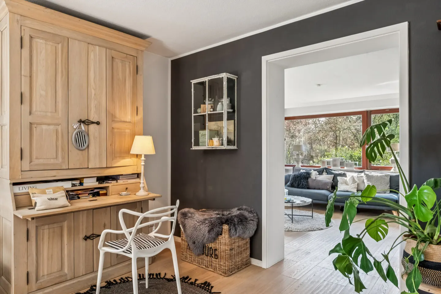 Interior view of a home office with a wooden desk, white chair, and a glimpse into a living room with a blue sofa.
