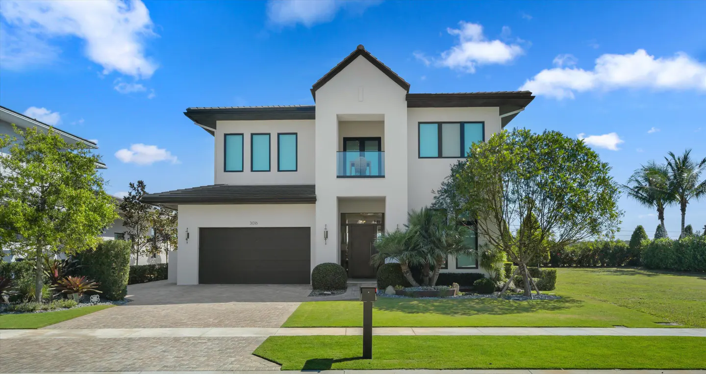 Two-story white house with a dark roof, a balcony, a dark garage door, and a green lawn on a sunny day.