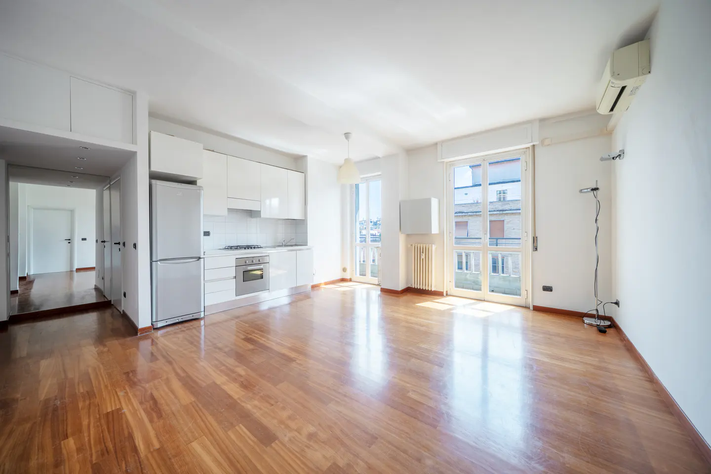 Bright, empty apartment with hardwood floors, white walls, and a modern white kitchen. Balcony doors let in natural light.