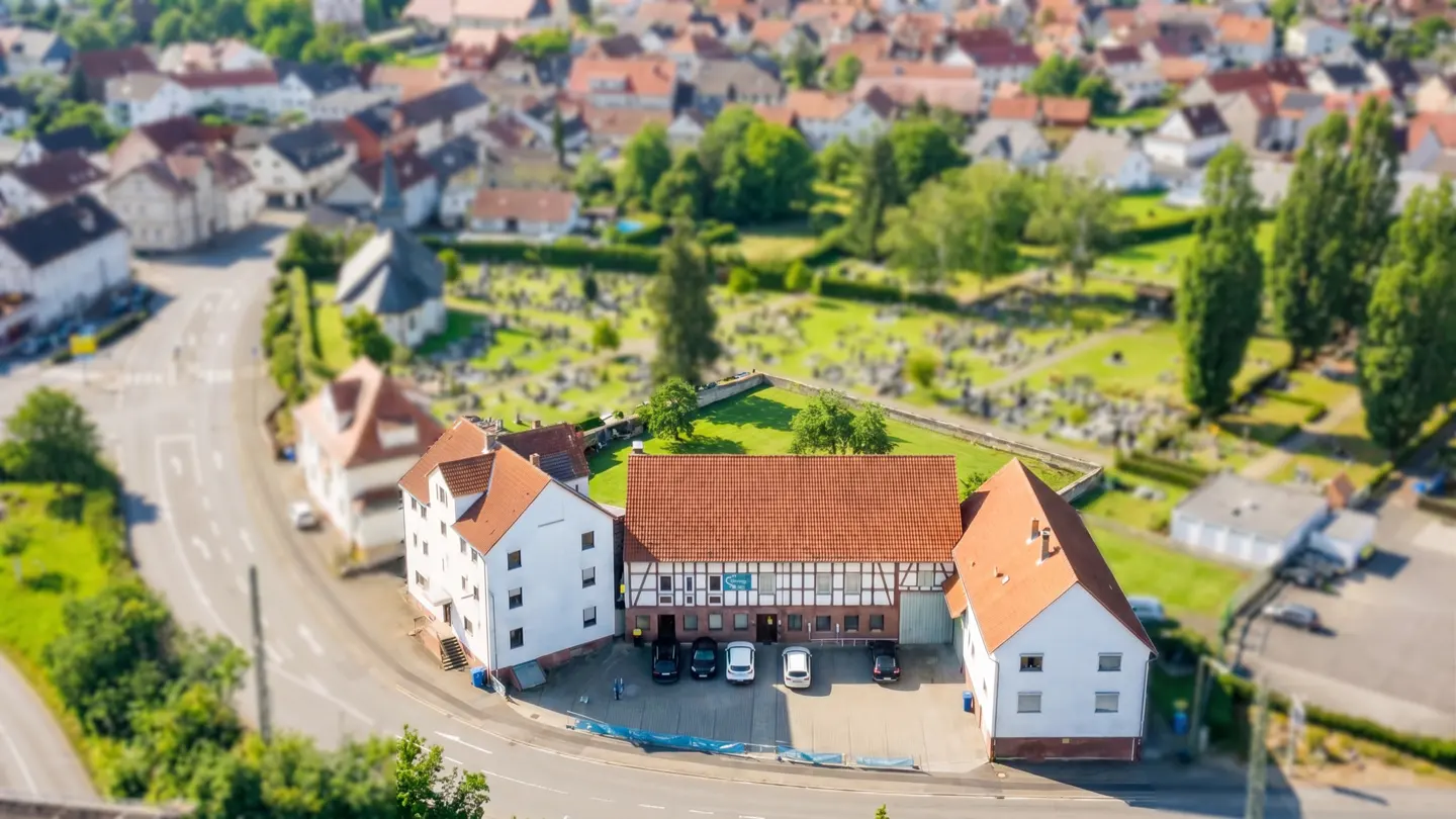 Aerial view of a white building with a red tile roof, surrounded by a town and cemetery. Cars are parked in front.