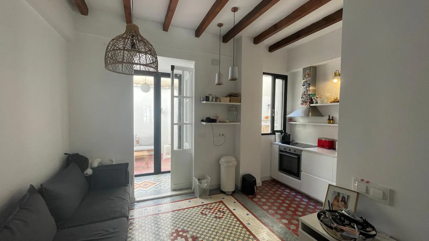 A bright, open-concept living space with a gray sofa, white walls, and a small kitchen area. The floor has decorative tiles, and a door leads to a small outdoor patio.