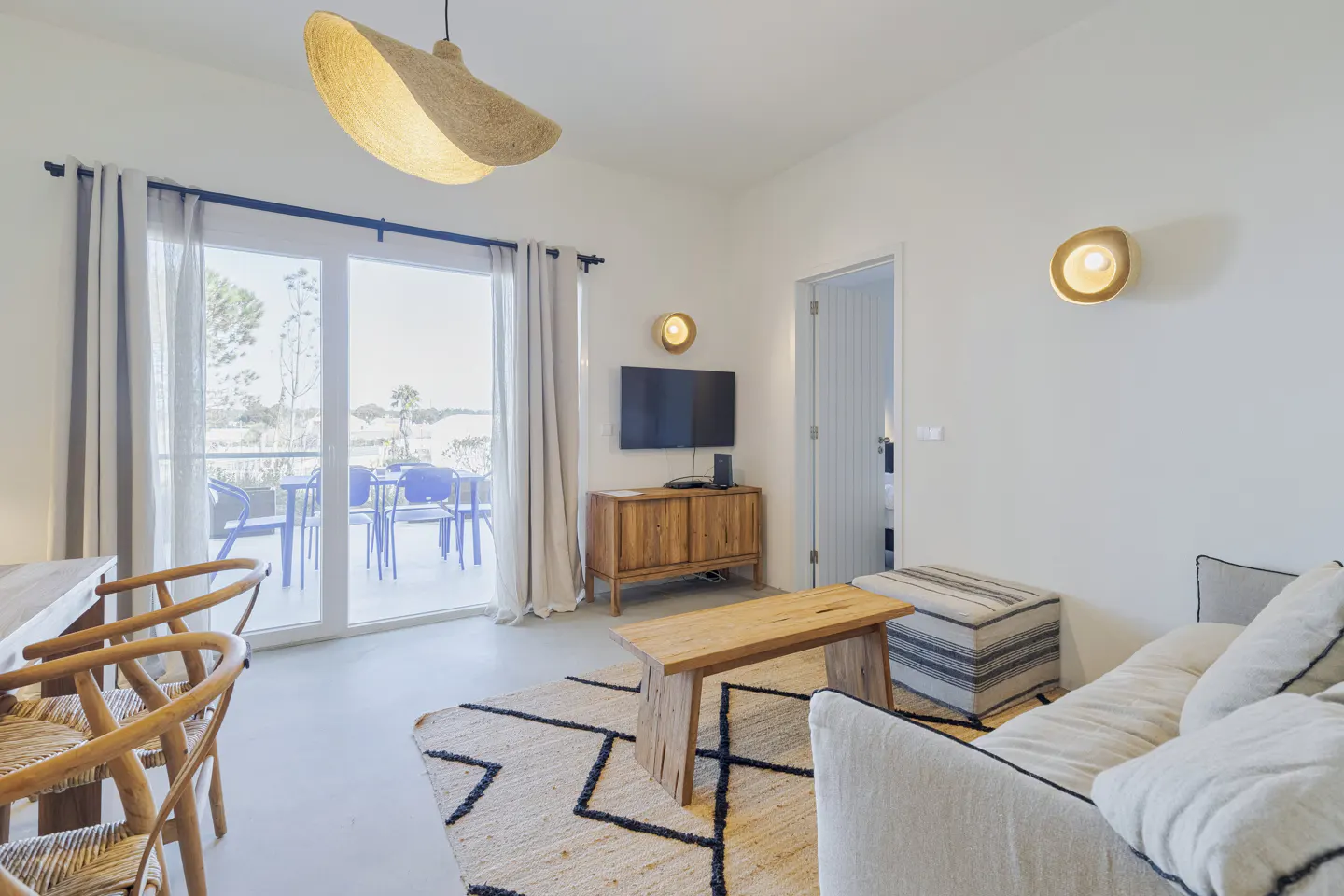 Bright living room with white walls, a wood TV stand, a beige sofa, and a jute rug with black geometric patterns. A patio is visible through glass doors.