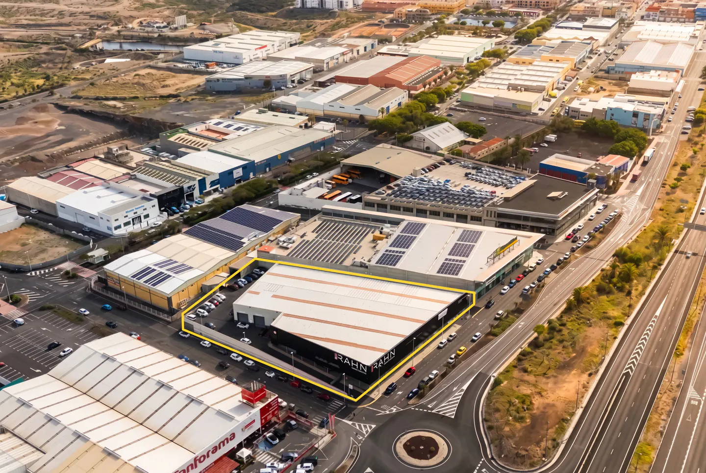 Aerial view of a large, modern, light gray commercial building with "RAHN" signage, outlined in yellow, surrounded by parking and other buildings.