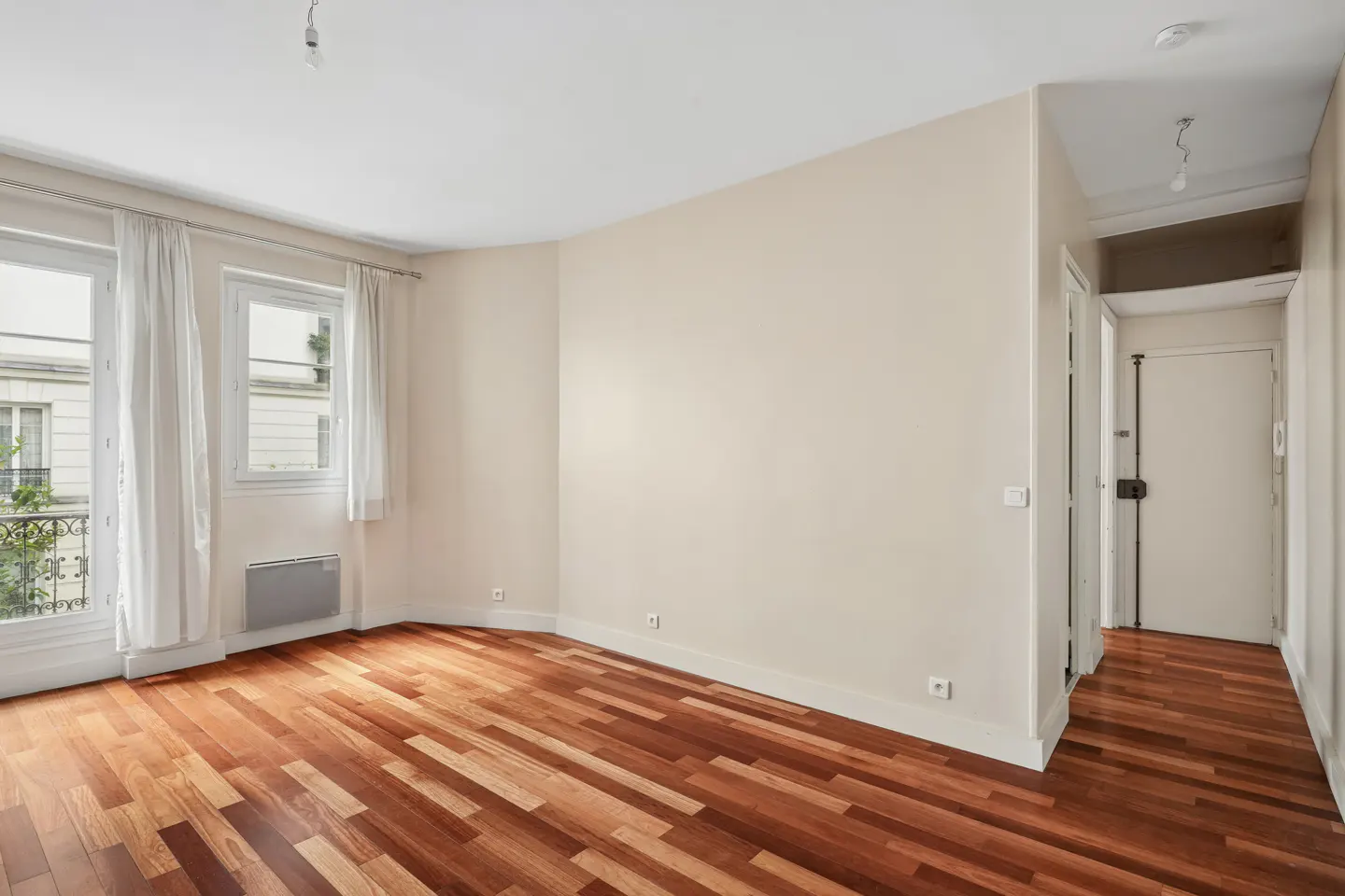 Bright, empty room with hardwood floors, cream walls, and white trim. Two windows with sheer curtains overlook a building. A hallway leads to a white door.