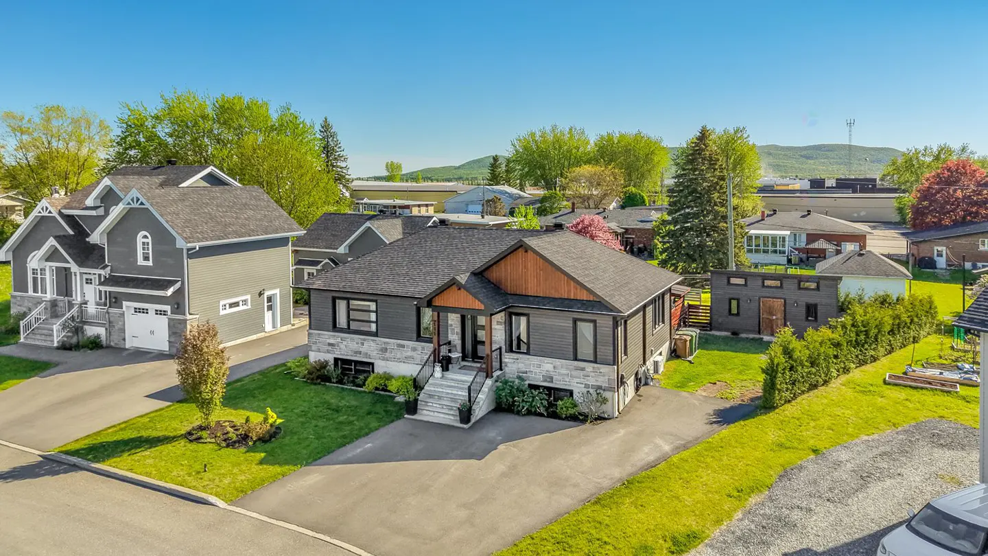Aerial view of a gray two-story house with stone accents, a dark roof, and a wood-trimmed entrance. Green trees and blue sky in the background.