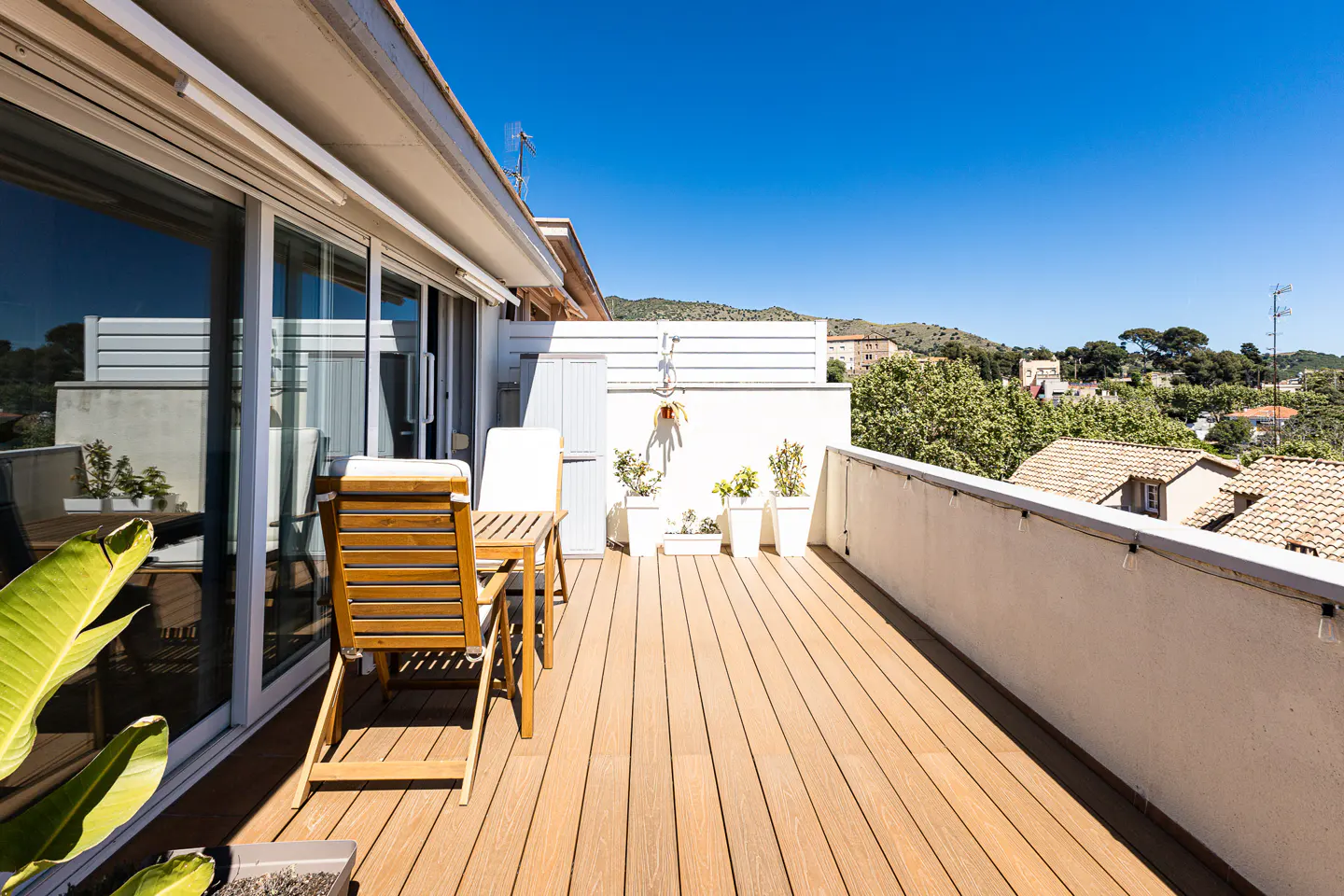 A sunny balcony with a wooden deck, table, chairs, and potted plants, overlooking a cityscape with trees and buildings.