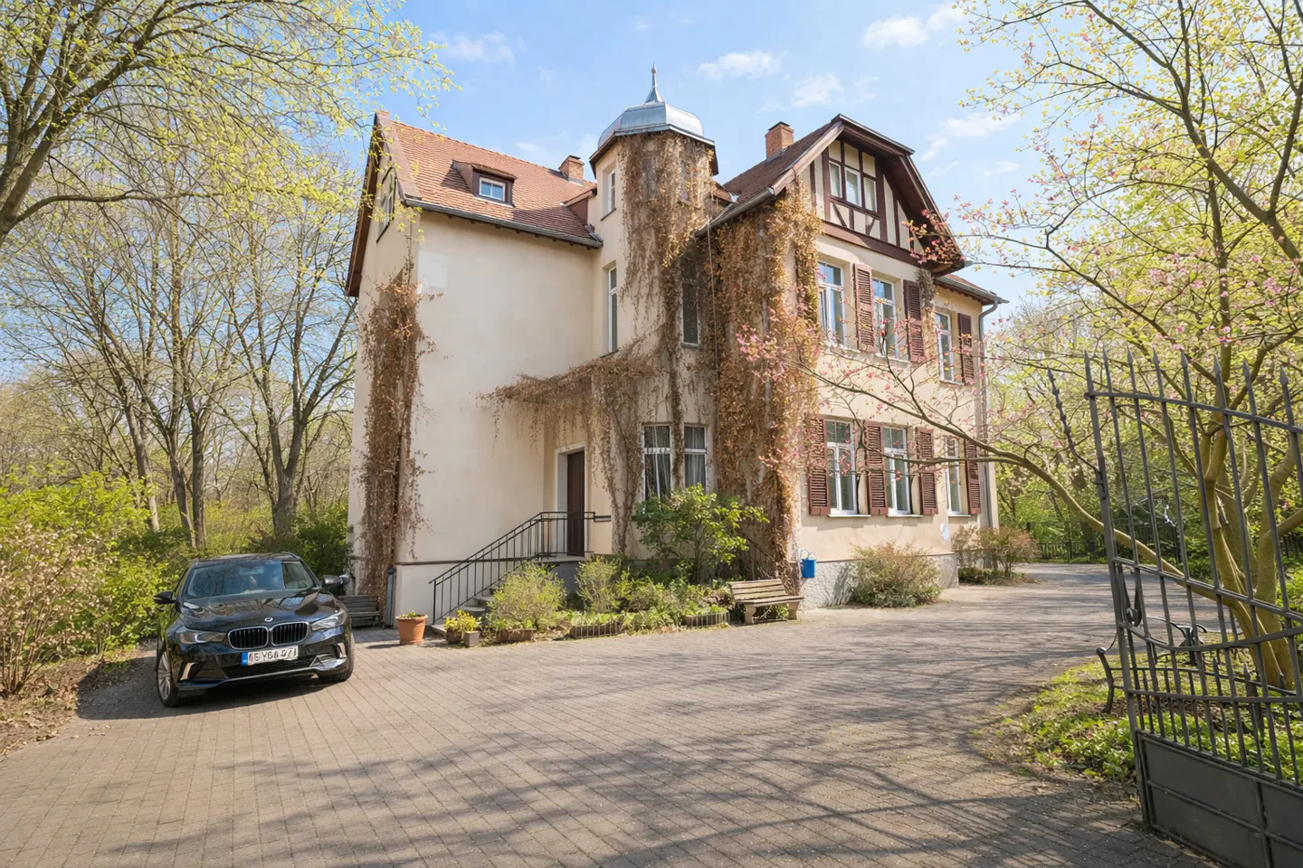 A three-story beige house with brown shutters and a black car parked in the driveway. Vines climb the walls.