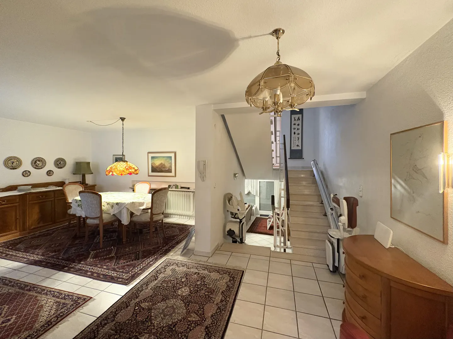 A bright, open-concept dining and entry area with a staircase and chair lift. The dining area has a table, chairs, and a wooden sideboard.