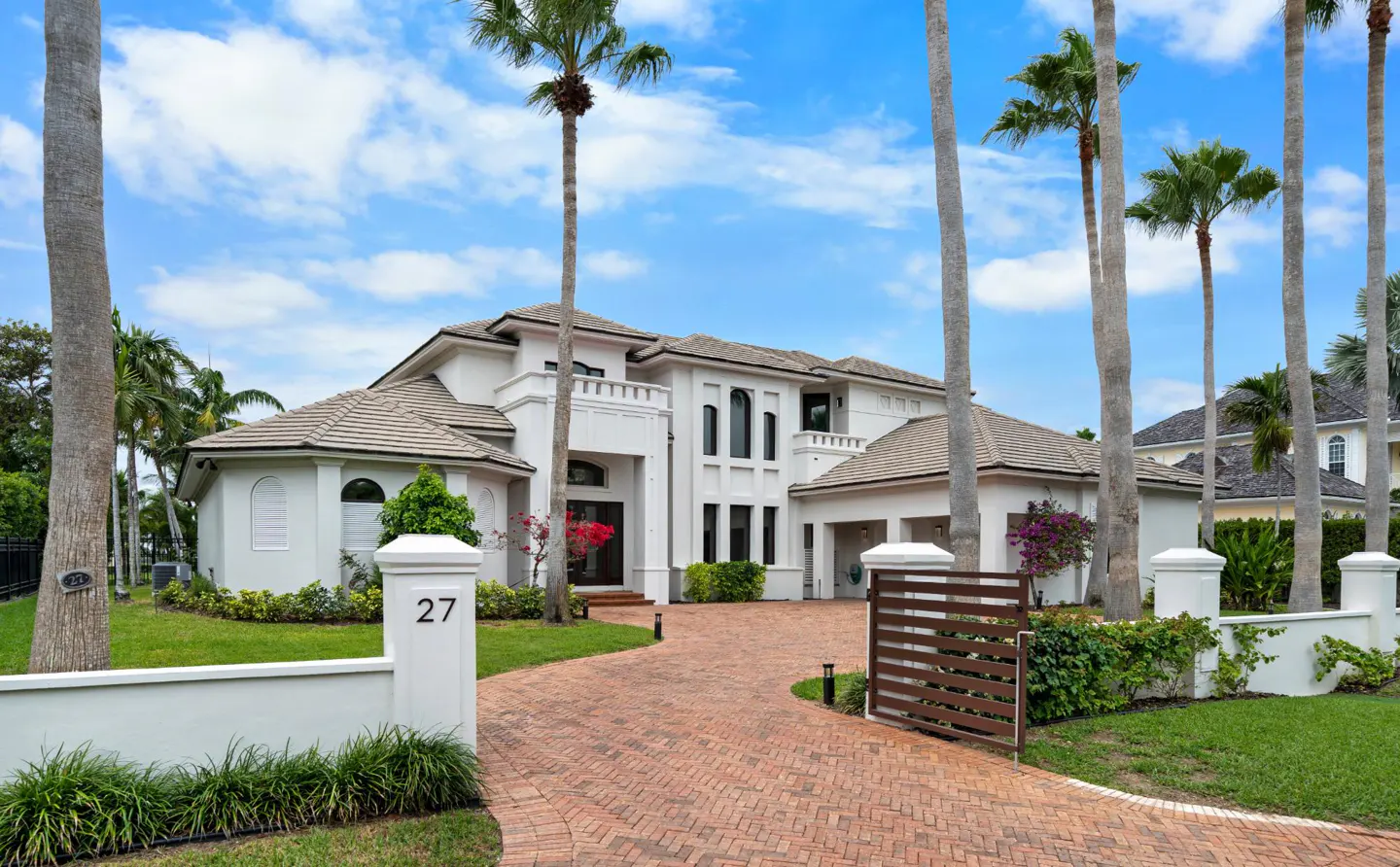 A large, white, two-story house with a brown tile roof, palm trees, and a brick driveway. The house number "27" is visible on a white pillar.
