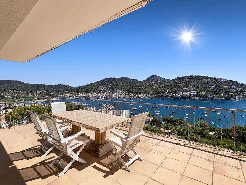 Outdoor patio with table and chairs overlooking a bay with boats, mountains, and a sunny blue sky.