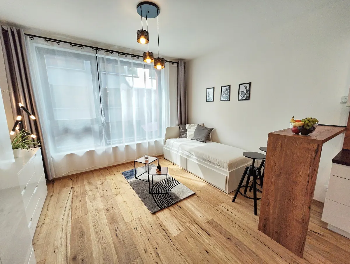 Bright studio apartment with wood floors, a daybed, and a breakfast bar with stools. Three framed prints hang on the wall.
