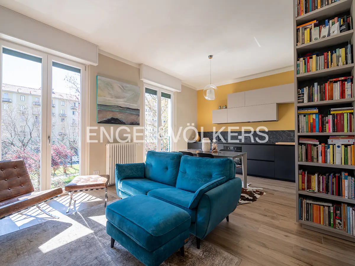 Bright living room with a blue sofa, brown leather chair, and a full bookshelf. Large windows overlook a tree-lined street.