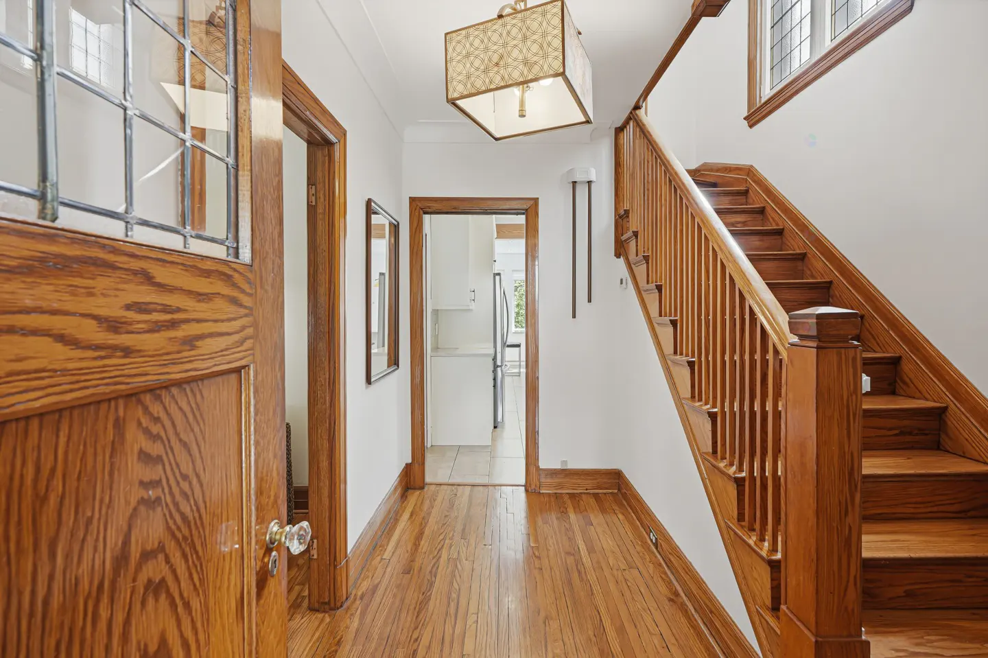 Bright entryway with hardwood floors, a wooden staircase, a decorative pendant light, and a view into a white kitchen.