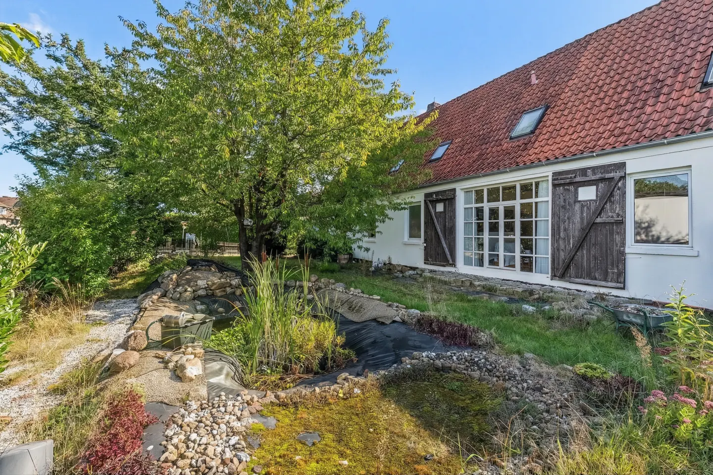 Exterior view of a white house with a red tile roof, a pond, and a large green tree.