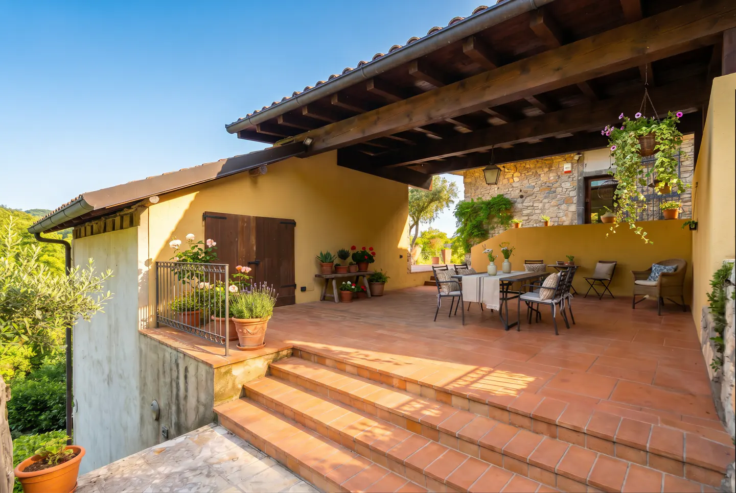 Spacious terracotta patio with outdoor dining set, potted plants, and a covered wooden pergola against a stone house wall.