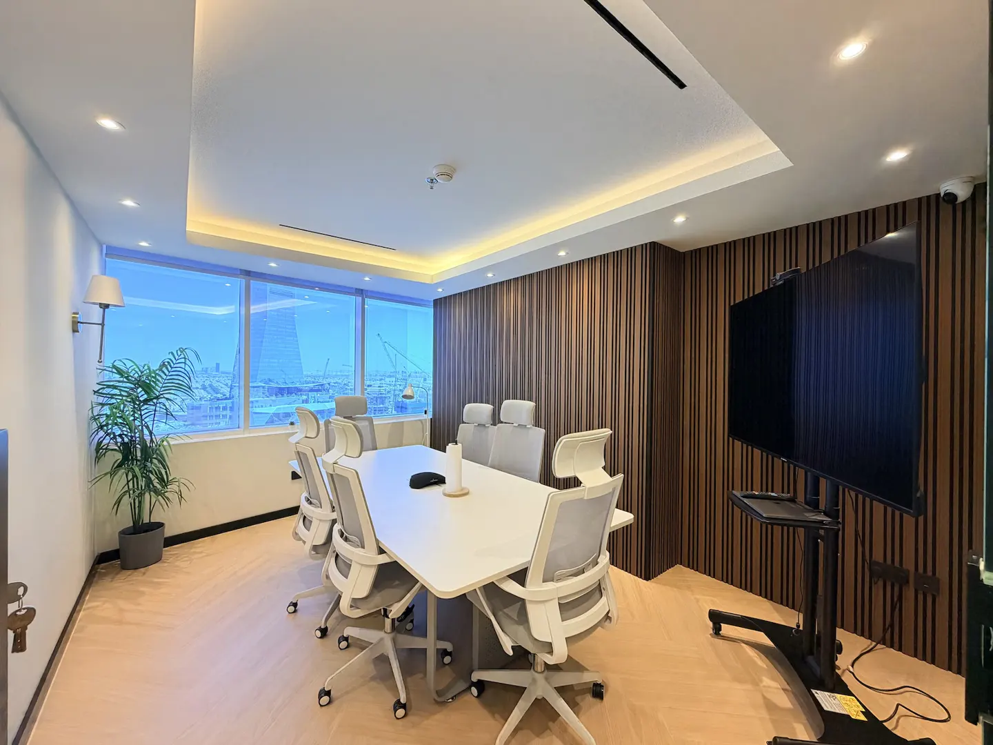 Modern conference room with a white table, grey ergonomic chairs, wood-slat wall, large TV, and city view through windows.