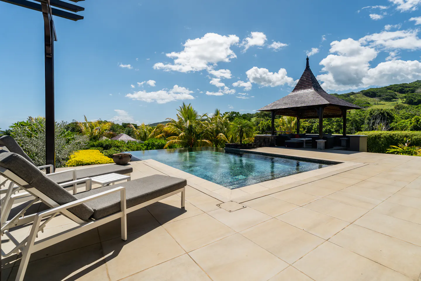 Luxury pool with lounge chairs, gazebo, and lush green landscape under a blue sky with clouds.