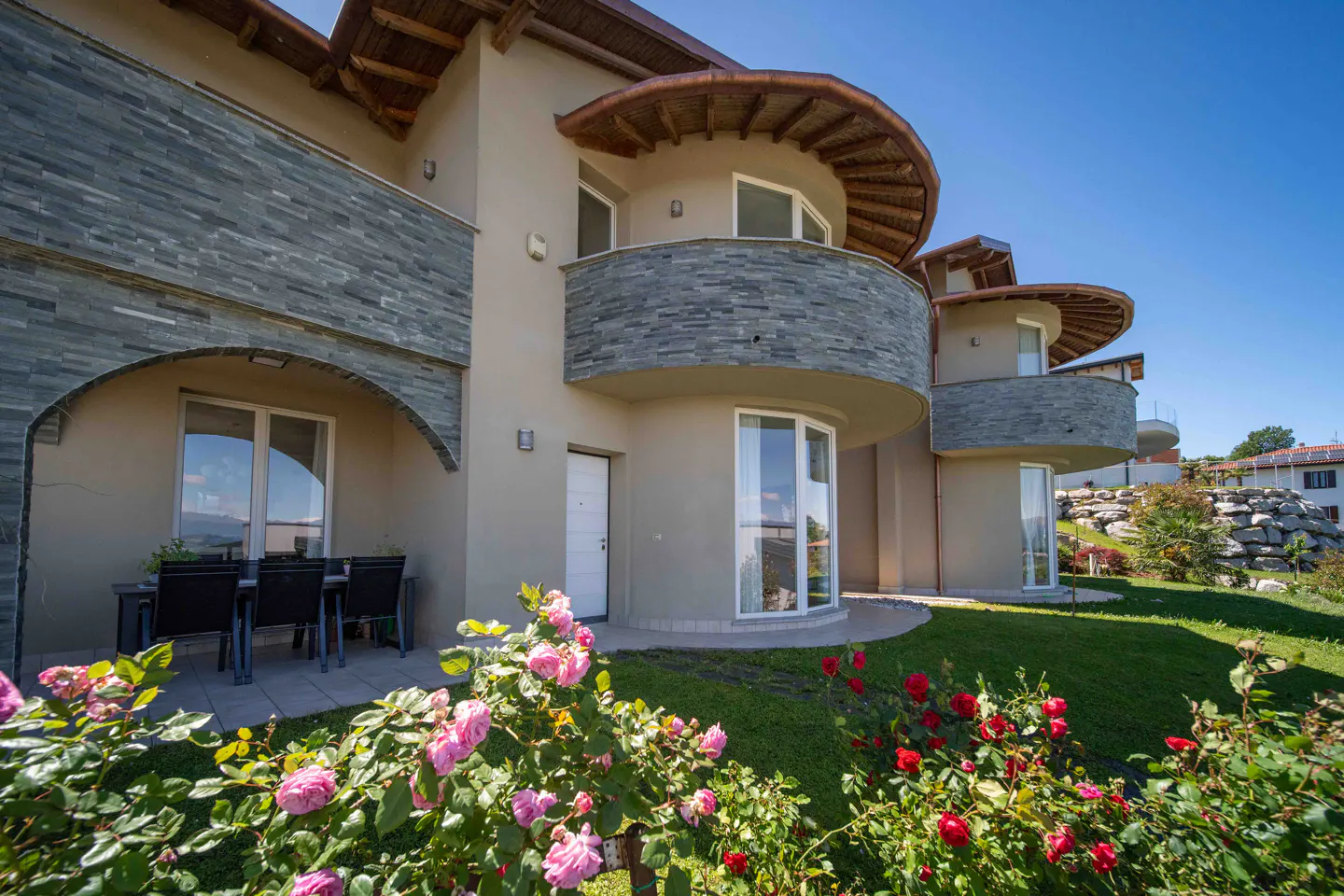 Exterior of a modern beige house with stone accents, round balconies, and a garden with pink and red roses.