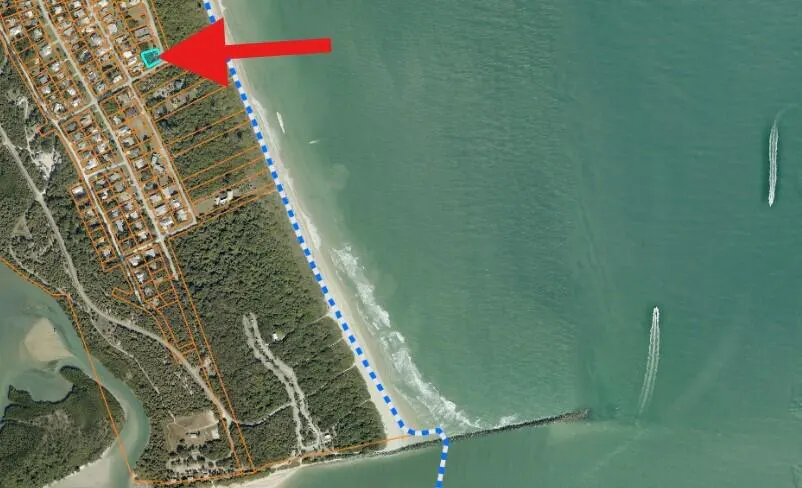 Aerial view of a coastal neighborhood with a red arrow pointing to a teal-outlined property near the beach. Boats are visible in the ocean.
