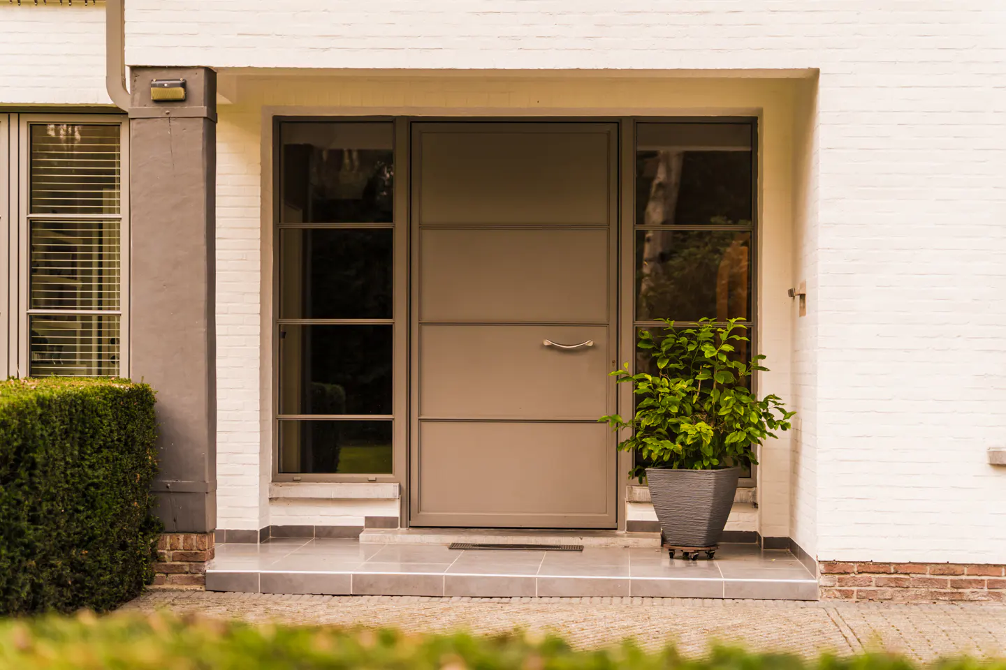 Front entrance of a white brick house with a taupe door and sidelight windows. A potted plant sits to the right of the door.