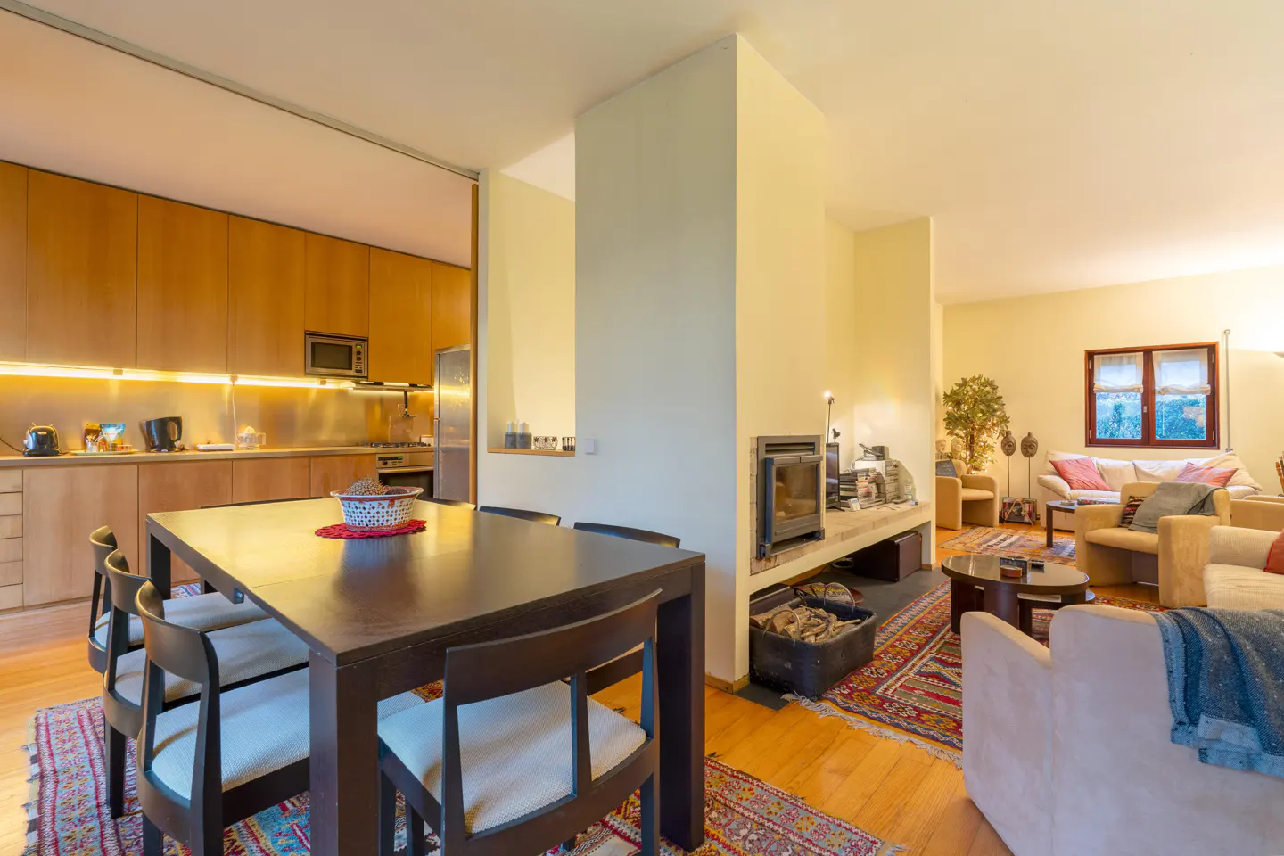 Open-concept living space with a dark wood dining table, chairs, and a kitchen with wood cabinets. A fireplace and living room are in the background.