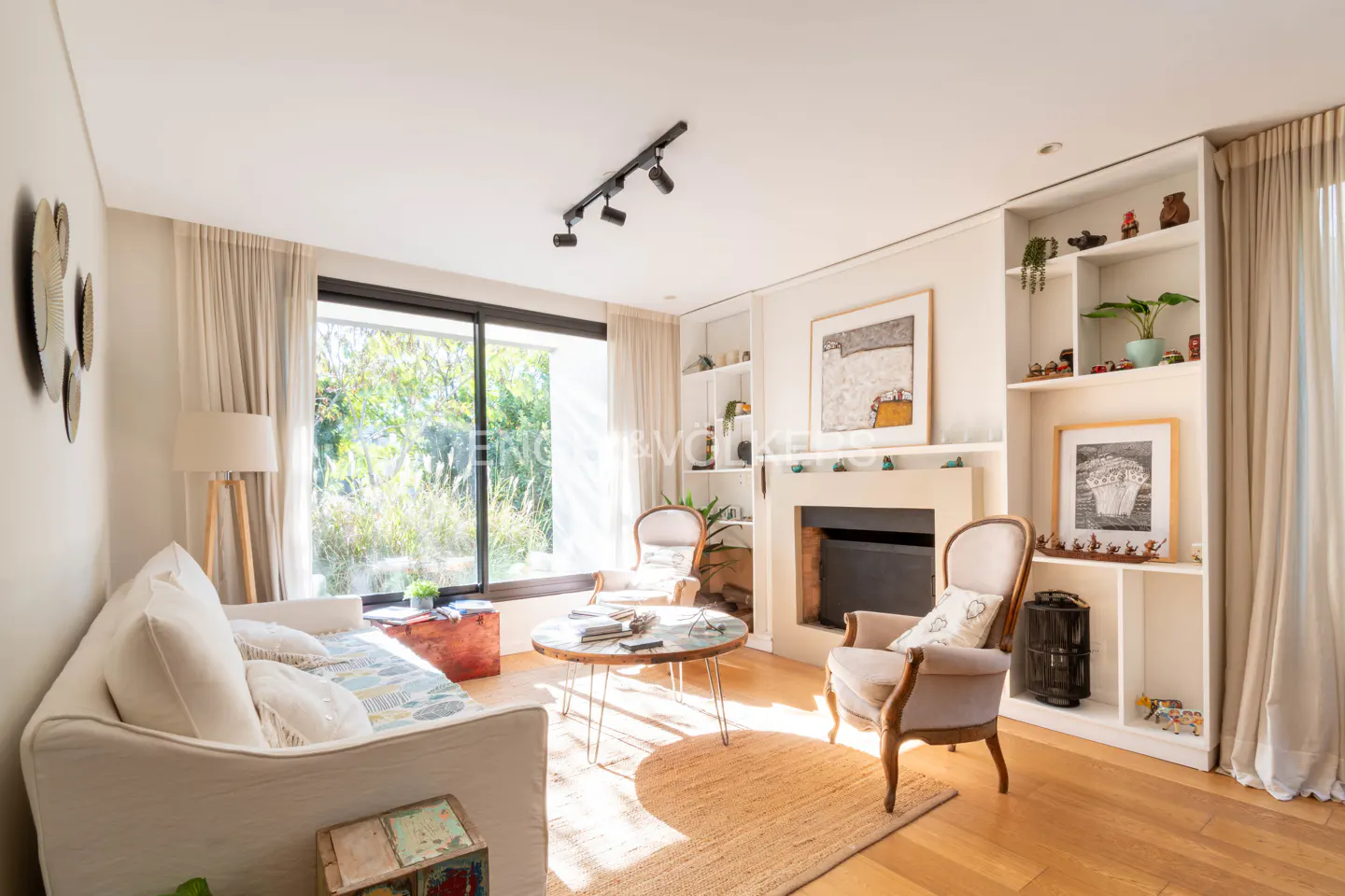 Bright living room with a white sofa, two armchairs, a fireplace, and built-in shelves. A large window overlooks a green garden.