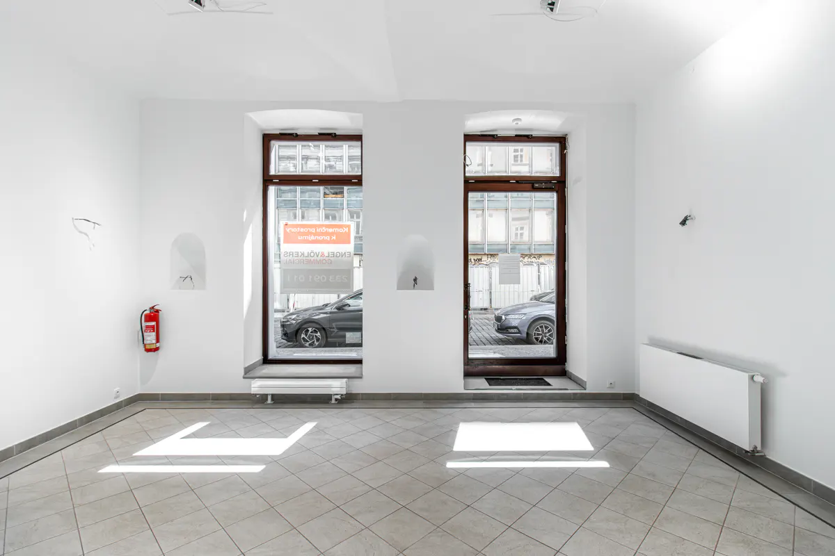 Empty white room with tile floor, two brown-framed windows showing street view with cars, and a red fire extinguisher on the wall.