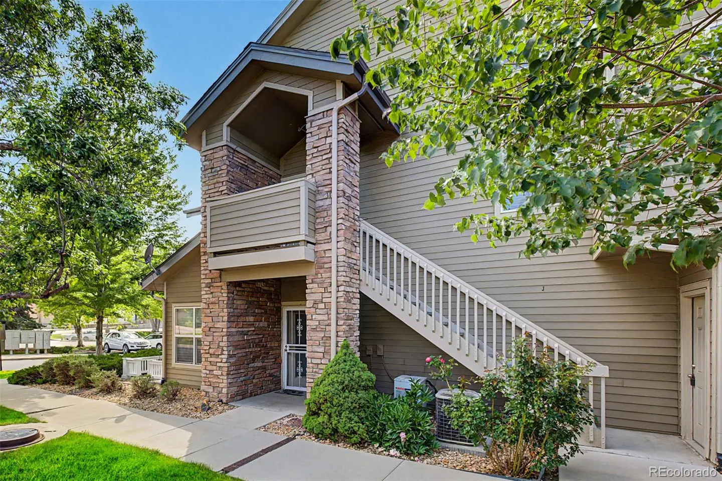 Exterior view of a two-story tan condo with stone accents, a small balcony, and a white staircase leading to the second floor.