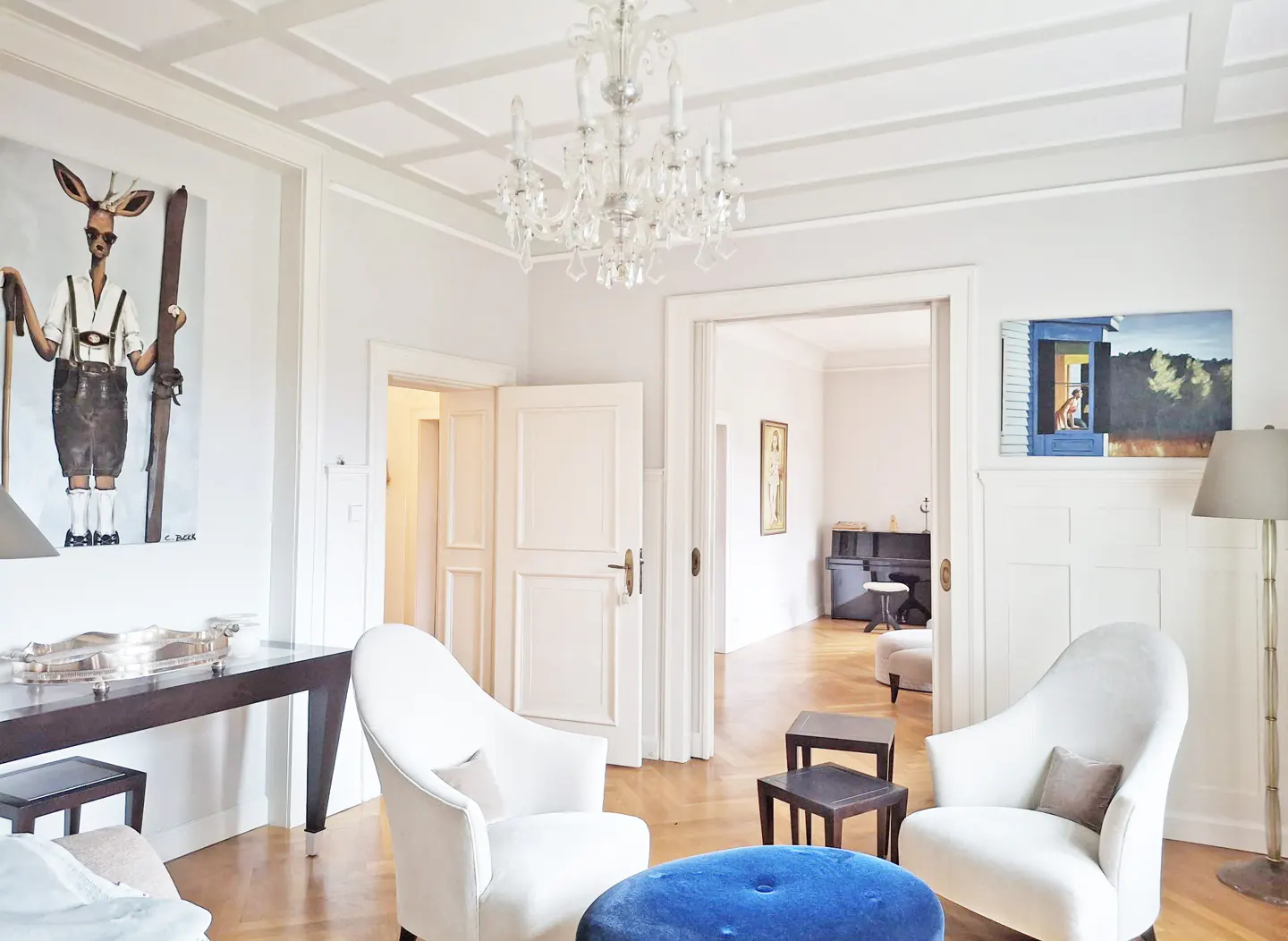Bright living room with white walls, wood floors, and coffered ceiling. A chandelier hangs above two white chairs and a blue ottoman. Artwork adorns the walls.