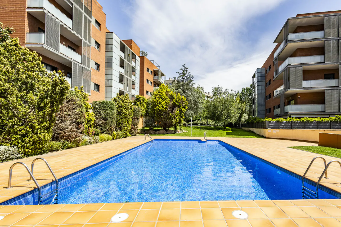 Outdoor pool with blue water and yellow tile surround, between brick apartment buildings and green trees under a blue sky.