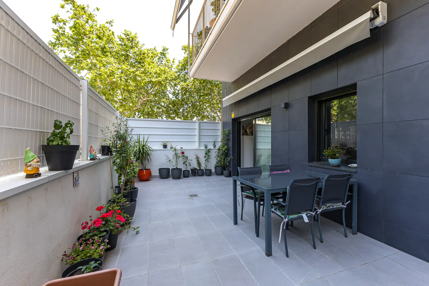 Outdoor patio with gray tile, a dark gray building, a table with four chairs, and potted plants.