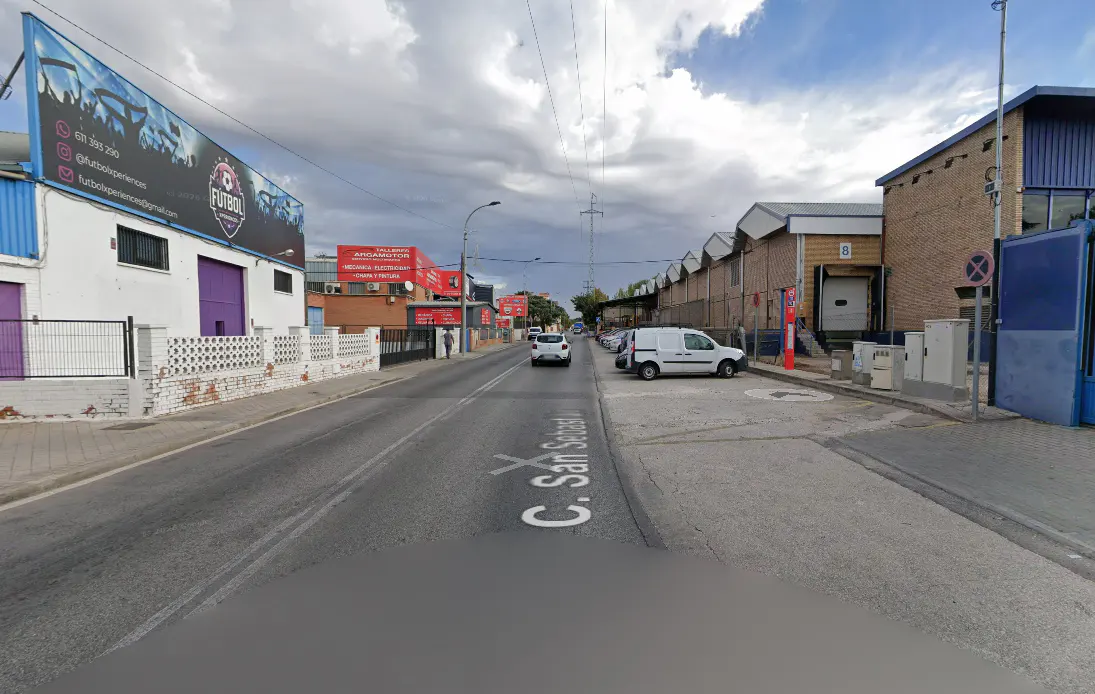 Street view of C. San Sebastian with warehouses and businesses under a cloudy sky. Cars and vans are parked along the road.