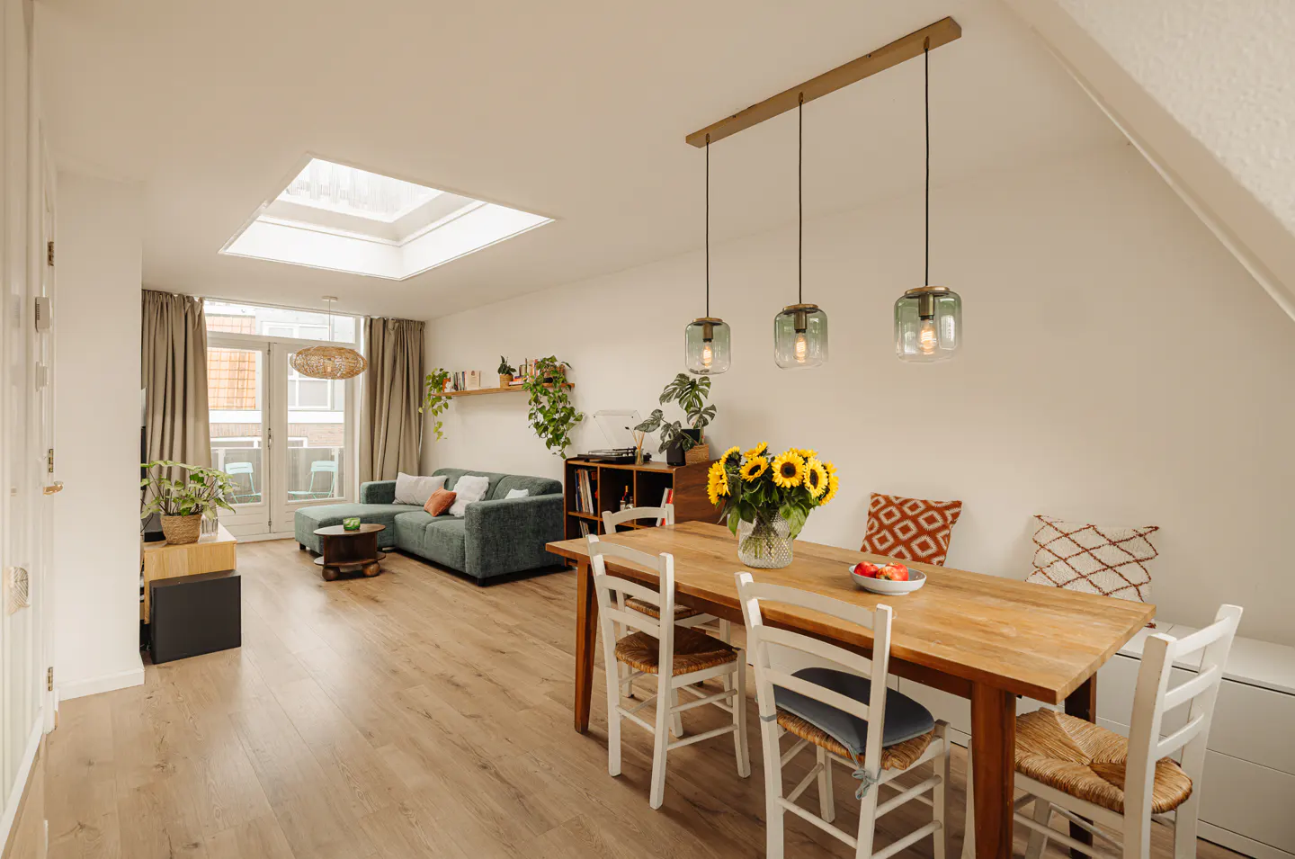 Bright, open living space with wood floors, a teal sofa, and a dining table with sunflowers. Skylight and pendant lights add natural and artificial light.