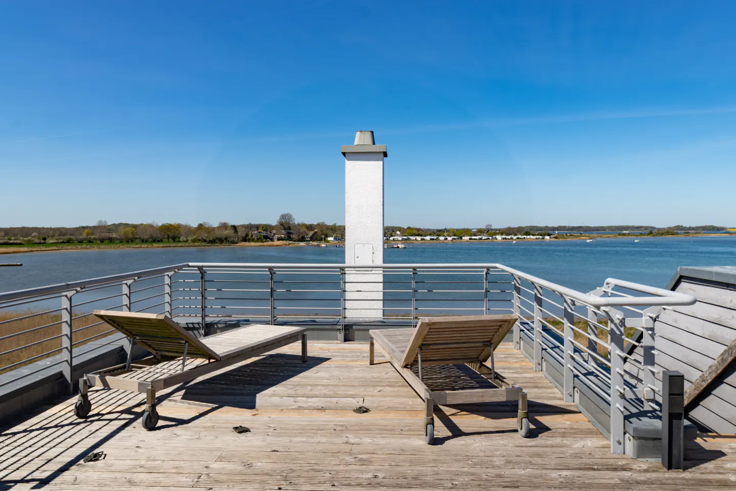 Wooden deck with two lounge chairs overlooking a blue bay. Metal railings surround the deck. A white chimney stands tall in the background.