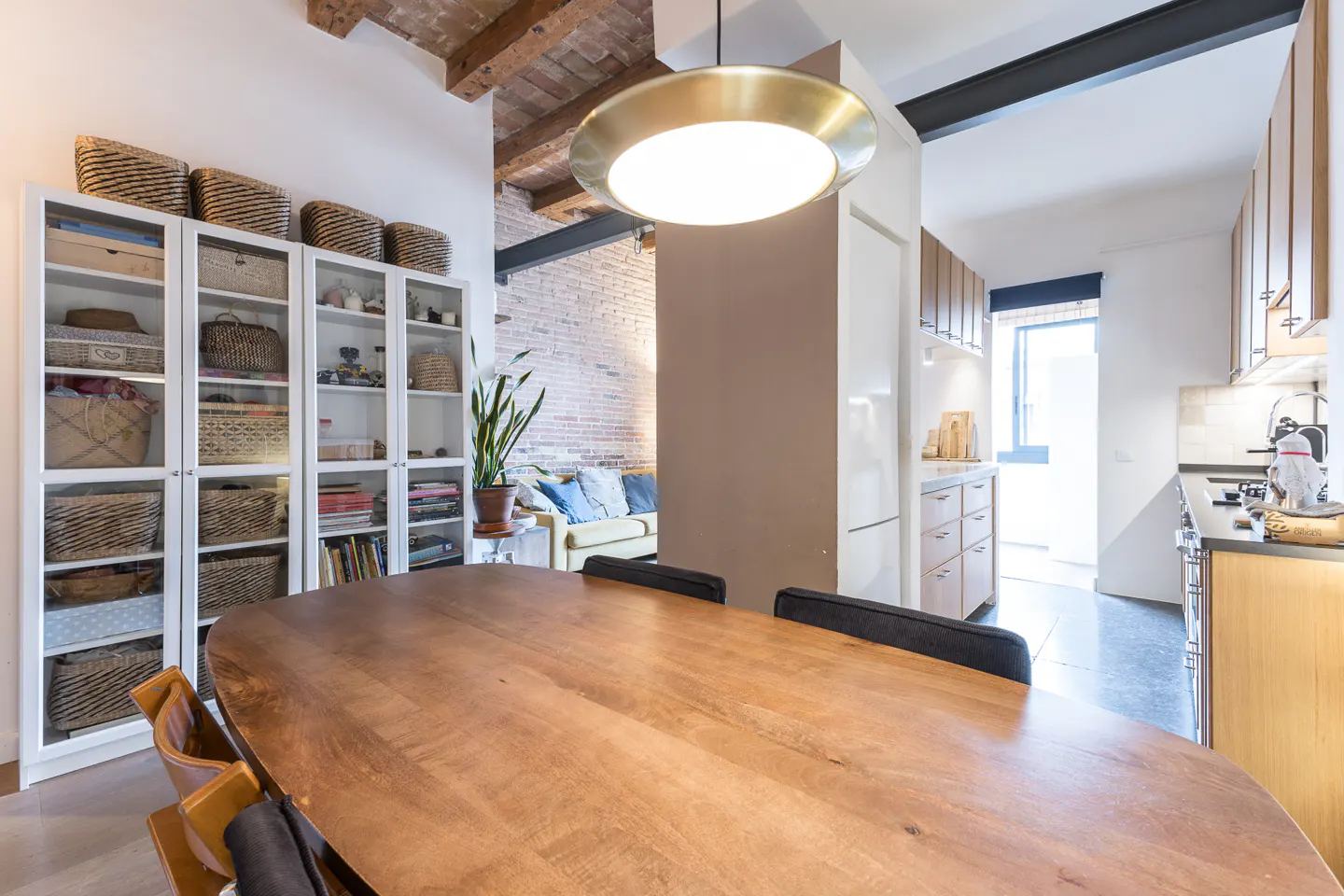 Modern dining area with a wooden table, glass-front storage cabinets, and a view into a kitchen with brick ceiling beams.