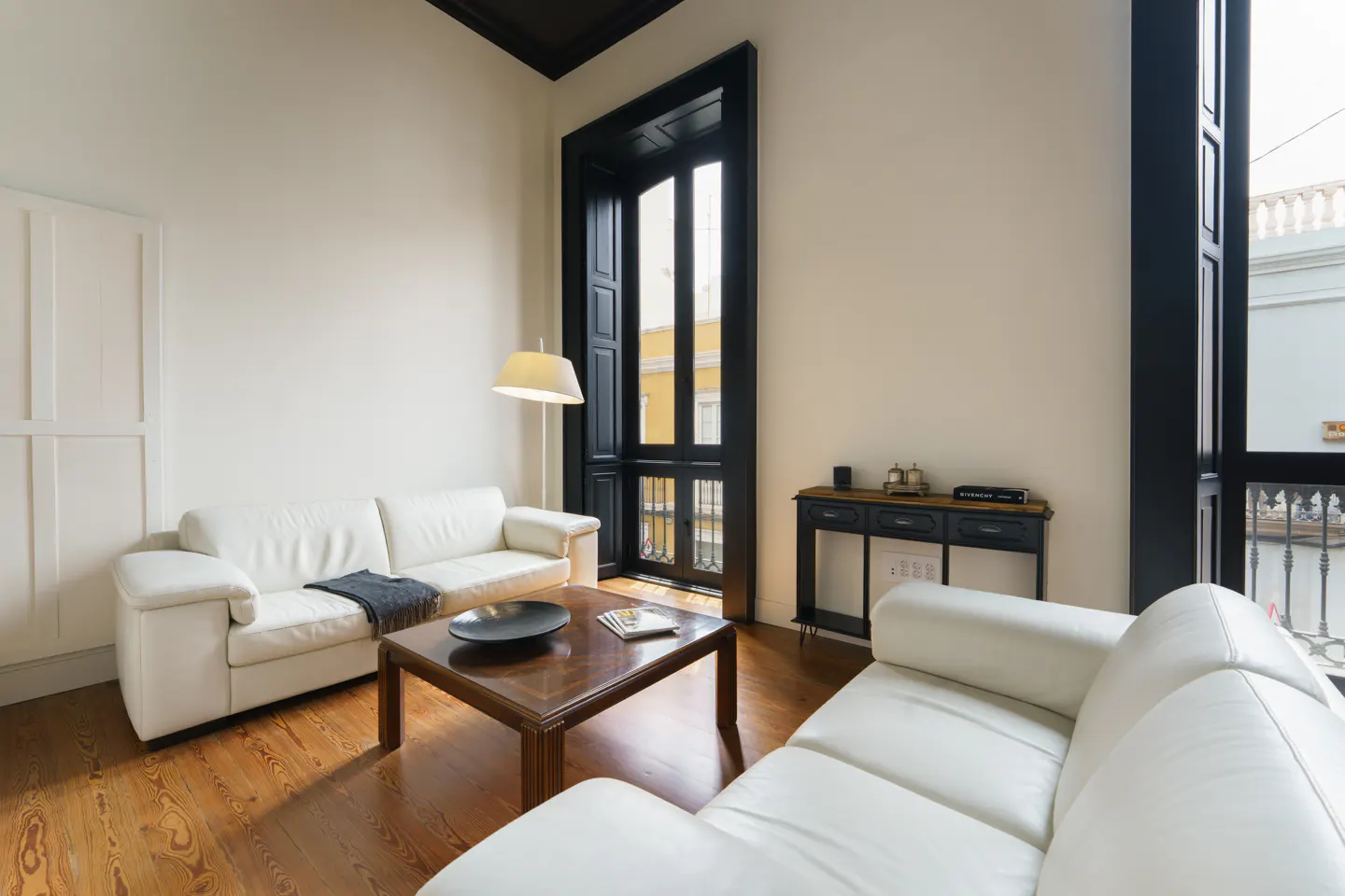 Bright living room with white walls, wood floors, and two white sofas. A dark wood coffee table sits in the center. Tall black framed windows let in natural light.