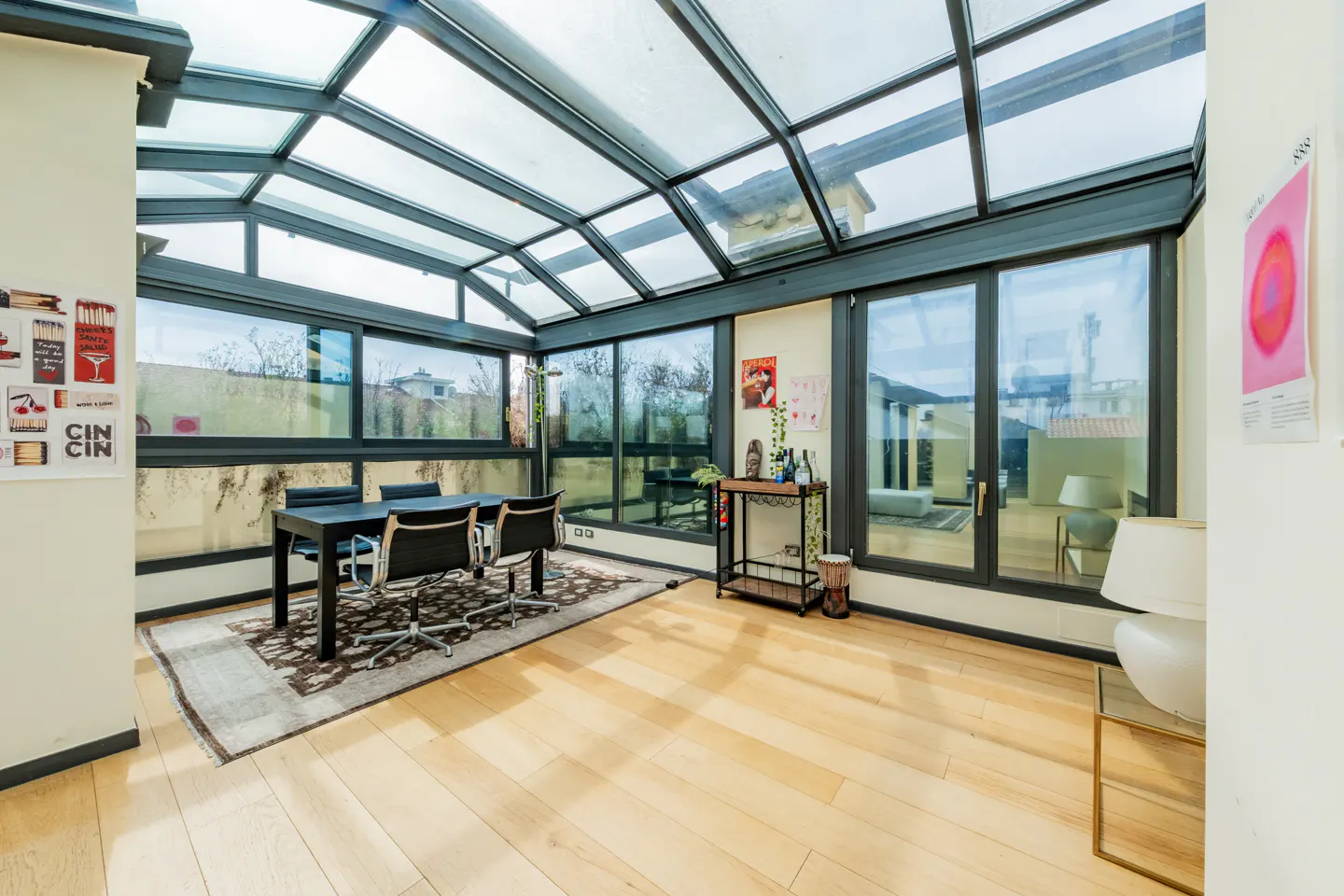 Bright sunroom with a glass ceiling, black table and chairs on a patterned rug, and light wood floors.