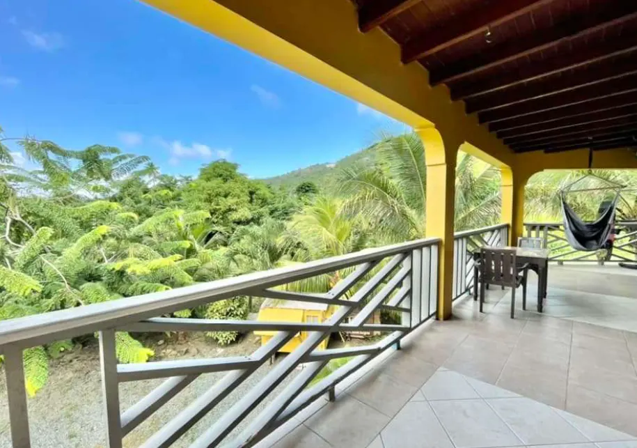 Balcony view with lush greenery. Yellow pillars frame the scene, with a table, chairs, and hammock visible on the tiled patio.