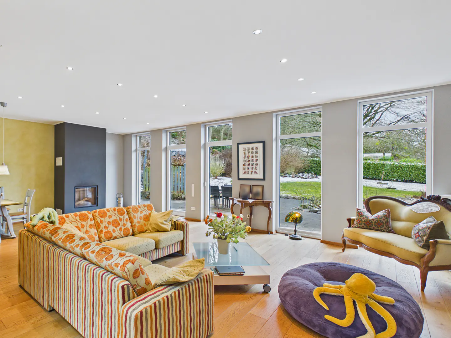 Bright living room with hardwood floors, large windows, and colorful sofas. A fireplace and garden view add to the ambiance.
