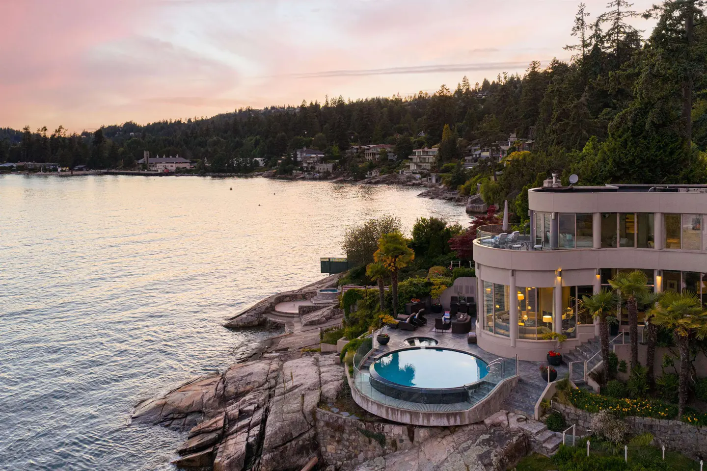 Modern waterfront home with a curved exterior, circular pool, and patio overlooking the ocean at sunset.