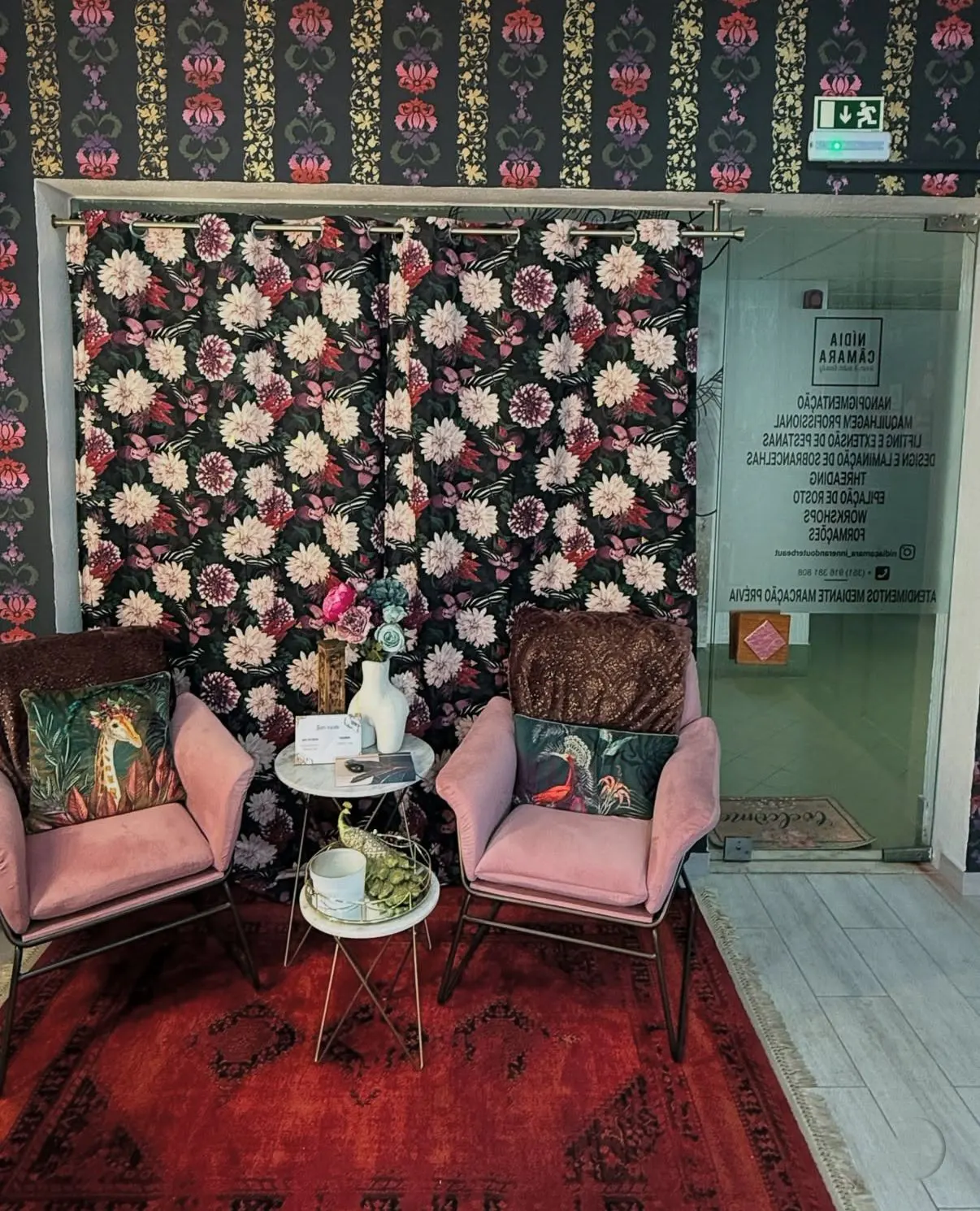 Cozy waiting area with two pink chairs, floral curtains, and a red rug. Tables with vases and decorative items add charm. A glass door is visible.