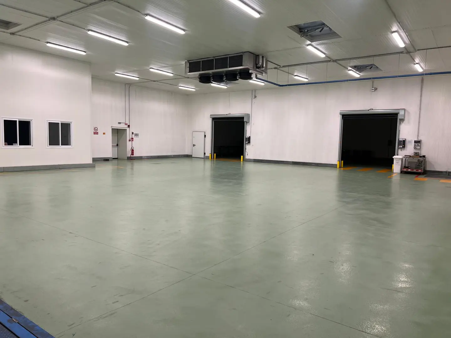 A large, empty warehouse with white walls, a light green floor, and two open loading dock doors. Overhead lighting and a cooling unit are visible.