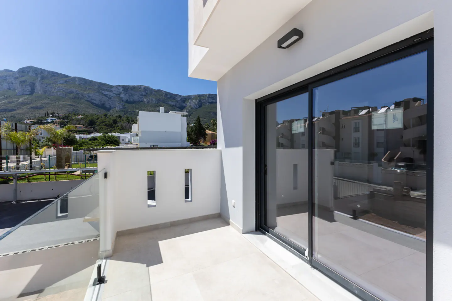 Modern white balcony with glass railings, large sliding glass doors, and a scenic view of mountains under a clear blue sky.