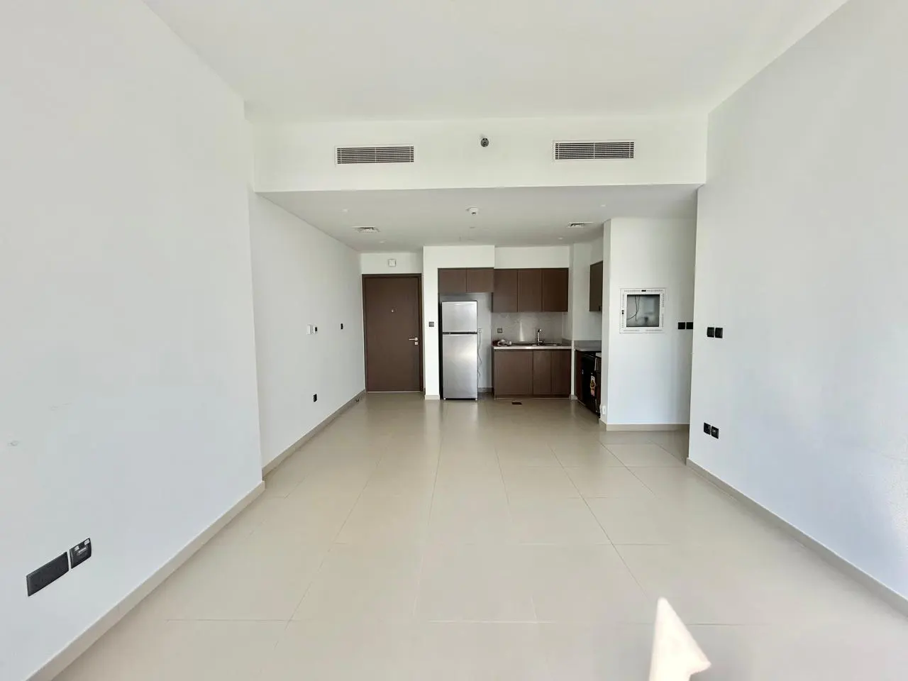 Bright, empty apartment with white walls and beige tile floor. A brown door and kitchen with stainless steel refrigerator are visible.
