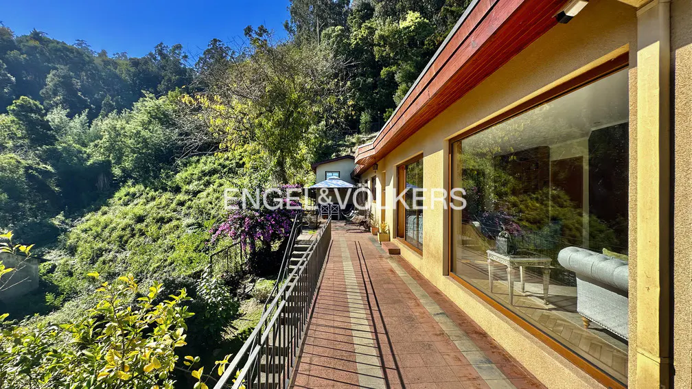 Exterior view of a tan house with a red brick patio and black railing, surrounded by lush green trees.